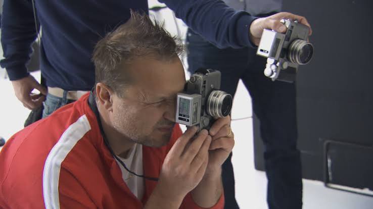 Juergen Teller taking a photo with a compact camera while someone behind him holds another one.